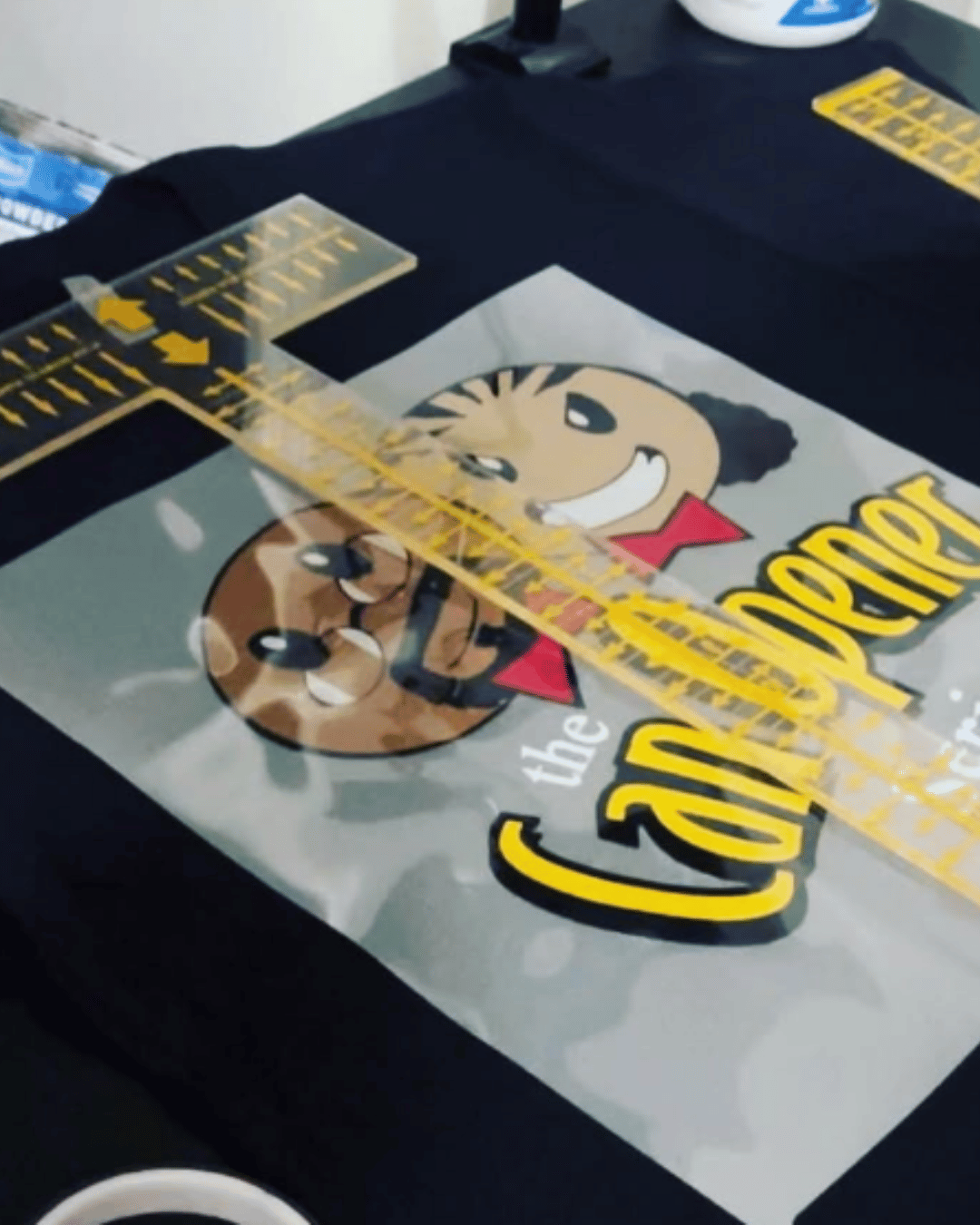 Printed transfer sheet with “The Can Opener” graphic placed on a black shirt during the custom apparel printing process.