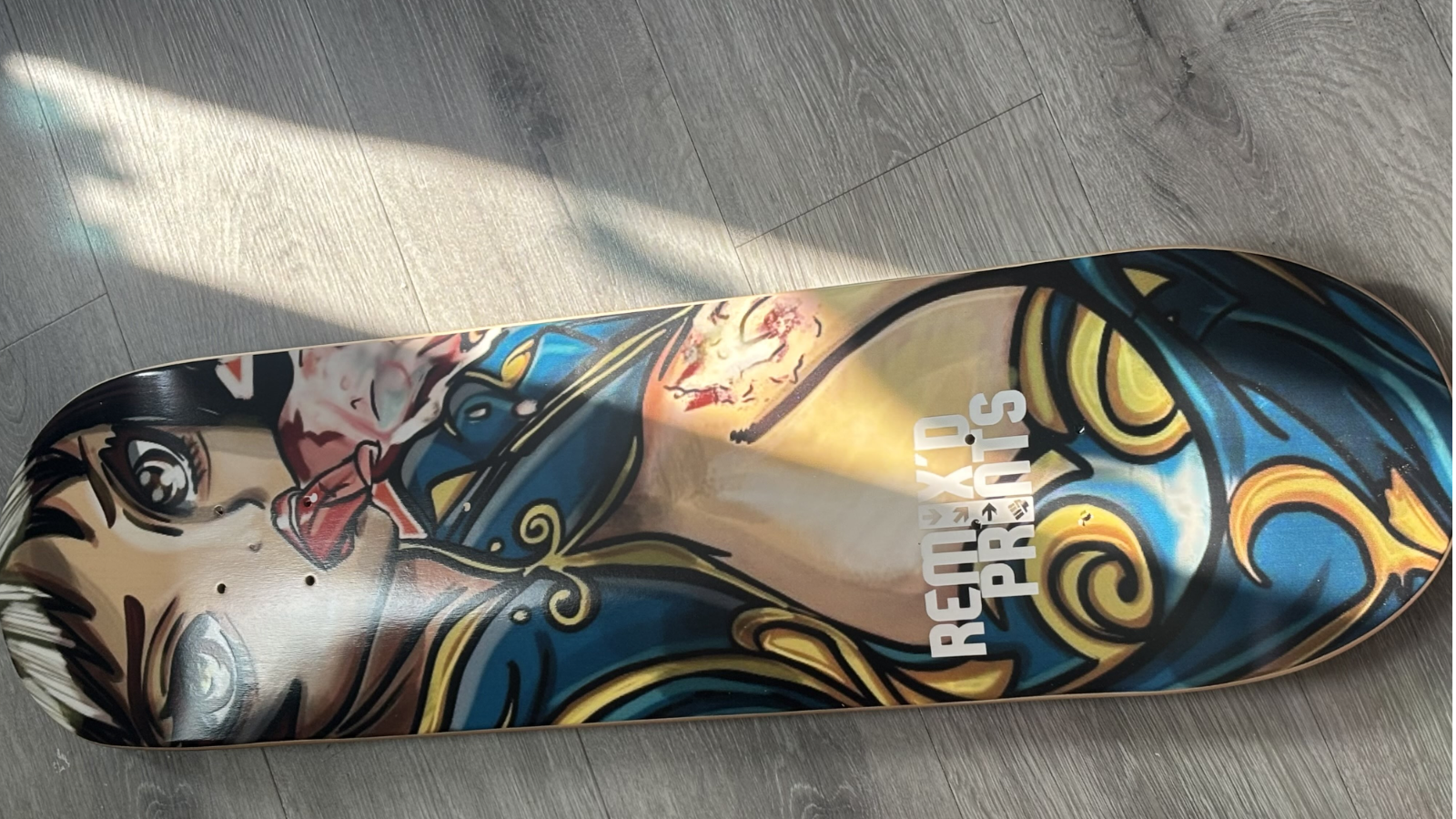 Skateboard deck featuring colorful illustrated artwork with flowing blue and gold elements and a vertical Remix’d Prints logo.